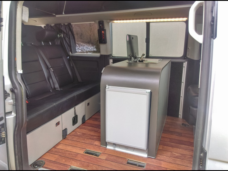 VW T5 – Aménagement sur mesure d'un salon central sur la base des sièges d'origine du Caravelle, avec îlot de cuisine/table au centre, équipé d'une plaque de cuisson CAN et d'un évier. Intégration d'un frigo Dometic.